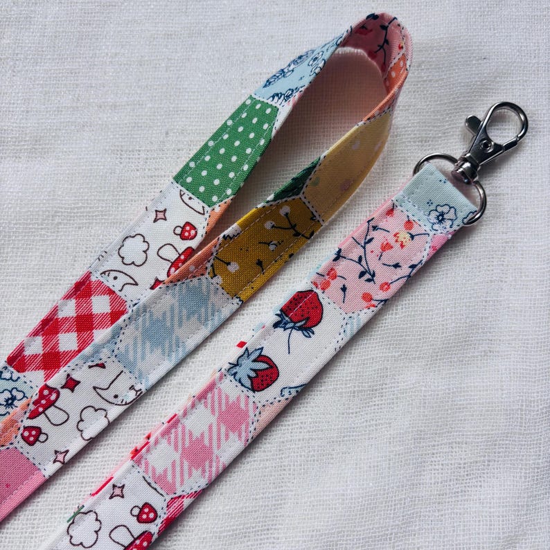 Op de afbeelding: Een stoffen lanyard met een patchworkontwerp van diverse patronen, waaronder rood-witte vichyruitjes, groene stippen, aardbeien, paddenstoelen en bloemmotieven. De band is bevestigd aan een zilverkleurige, draaibare metalen karabijnhaak.