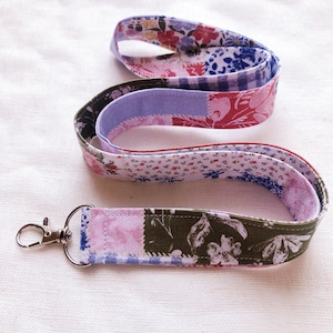 May include: A floral patterned lanyard with a silver-colored clasp. The fabric features a patchwork design with various floral prints in shades of pink, blue, and green. The lanyard is designed to be worn around the neck.