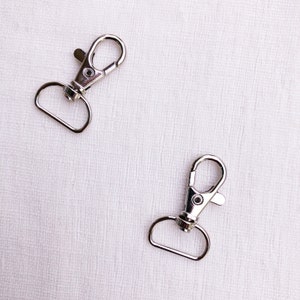 Op de afbeelding: Twee zilverkleurige metalen karabijnsluitingen, elk voorzien van een veerbelaste sluiting en een D-ring bevestigingspunt aan de basis. Deze hardware-onderdelen zijn ontworpen voor het verbinden van riemen of accessoires met een gladde afwerking.