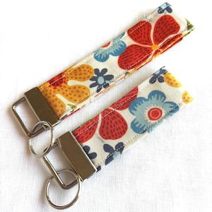May include: Two fabric key fobs with floral patterns. The key fobs have a cream background with red, blue, and yellow flowers. Each fob has a silver-colored metal clasp and a key ring.