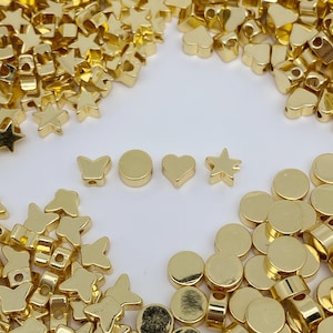 May include: A selection of gold-coloured metal beads in various shapes, including stars, hearts, butterflies, and circles. The beads have small holes for threading.