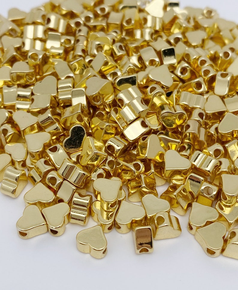 May include: A large pile of gold heart-shaped metal beads. The beads have a shiny finish and are ideal for jewellery making or crafting.