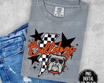 May include: A gray t-shirt with a graphic design of a bulldog wearing sunglasses and a checkered lightning bolt. The text "Bulldogs" is in orange lettering with black outlines. The design is surrounded by black stars.