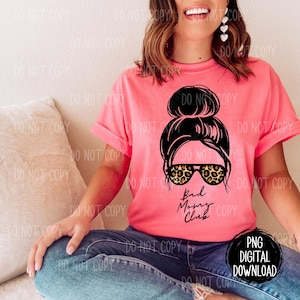 Puede incluir: Una camiseta rosa con una ilustración en blanco y negro de una mujer con un moño desordenado y gafas de sol de estampado de leopardo. El texto "Bad Moms Club" está impreso debajo de la ilustración.