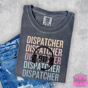 May include: A dark gray Comfort Colors t-shirt with the word "DISPATCHER" in a stacked design with a headset graphic and the number "911". The shirt is paired with blue denim shorts.