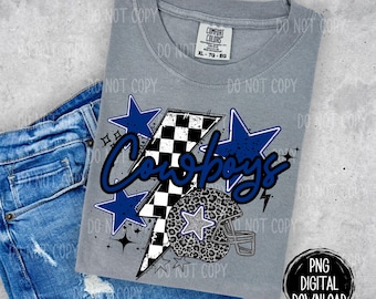 May include: A gray t-shirt with a graphic design featuring a football helmet with a leopard print design, a lightning bolt with a checkered pattern, and the word "Cowboys" in blue and white lettering. The design also includes blue stars.