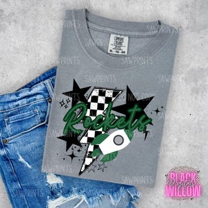 Może przedstawiać: Szary T-shirt z zielono-białą rakietą w kratkę i napisem "Rockets" w zielono-białym kolorze. Grafika otoczona jest białymi gwiazdami.