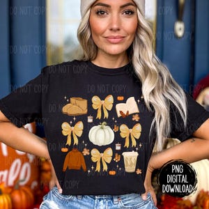 May include: A black t-shirt with a fall-themed design featuring a pumpkin, boots, a book, a cup of coffee, and fall leaves. The design is in shades of brown, orange, and yellow.