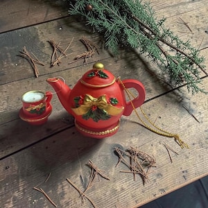 May include: A red teapot ornament with a gold bow and green holly leaves, featuring a matching miniature teacup ornament attached by a string. The teapot has a gold-colored lid knob and a gold beaded base, with a gold hanging cord attached to the handle.