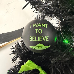 May include: A matte black spherical Christmas ornament featuring the neon green text I WANT TO BELIEVE printed above a graphic of a flying saucer. The ornament has a silver-toned metal cap at the top and is hanging from a black artificial tree.
