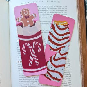 May include: Two pink bookmarks with festive designs. One features a gingerbread man in a red mug with marshmallows and candy canes. The other depicts a layered cake with red and white stripes. Both are set against an open book.