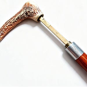 Pode incluir: Um cabo de bengala feito de material de chifre texturizado em tons de castanho e creme, fixado a um eixo de latão com marcas de medição métricas visíveis em centímetros, ligado a um colar prateado polido e a um eixo de madeira castanho-avermelhado.