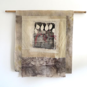 Puede incluir: Colgante de pared textil con una estética natural y terrosa. La obra de arte presenta un cuadrado central con un diseño de hojas en blanco y negro, acentuado con hilo rojo. La pieza está montada sobre una varilla de madera, mostrando una composición texturizada y en capas.