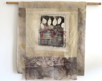 May include: A rectangular fabric wall hanging suspended from a wooden rod. The piece features layered, earth-toned textile panels with visible stitching. A central square patch displays a black and white print of leaves with red embroidered accents.