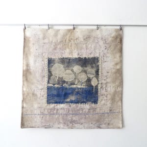 Op de afbeelding: Een vierkante textiele wandhanger met een centraal rechthoekig paneel met een donkerblauwe achtergrond en witte cirkelvormige boomachtige figuren. De omringende stof is gevlekt beige met gestikte randen en horizontale lijnen blauw borduurwerk.
