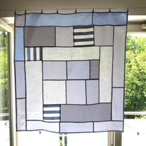 Op de afbeelding: Een vierkante patchwork quilt met een verscheidenheid aan blauwe en witte stoffen panelen. Het ontwerp bestaat uit rechthoekige en vierkante secties met afwisselende patronen van dunne strepen, dikke strepen en effen lichtblauwe of witte texturen.