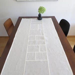 May include: A rectangular, dark brown wooden dining table with a white linen table runner. The runner has a brick-like pattern in the center. A small black vase with a green flower arrangement sits on the table. Two chairs are visible.