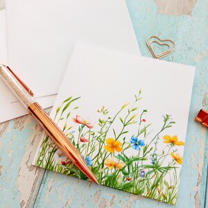 Puede incluir: Una tarjeta de felicitación cuadrada de color blanco con una ilustración en acuarela de flores silvestres coloridas en tonos amarillos, azules y rosas con tallos verdes. Un bolígrafo de oro rosa con cuerpo transparente y cristales descansa sobre ella.