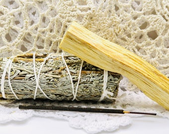 May include: A bundle of sage smudge sticks tied with white twine, a stick of palo santo wood, and a black incense stick on a white crocheted doily.