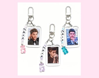 May include: Three clear acrylic keychains with a photo of a man in each. Each keychain has a beaded charm with the word "ZAYN" spelled out in white letters.