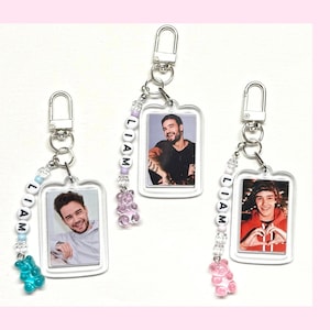 May include: Three clear acrylic keychain charms with a photo of a person inside each charm. Each charm has a silver key ring and a beaded chain with the name "LIAM" spelled out in white letters. The charms are decorated with a gummy bear charm in pink, blue, and purple.