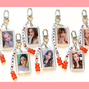 May include: Seven clear plastic keychains with red and white beaded chains. Each keychain has a small rectangular photo card with a different member of a girl group. The photo cards have white borders and the names of the members are spelled out on the chains: Handong, Siyeon, Yoohyeon, Gahyun, Dami, Jiu, and Sua.