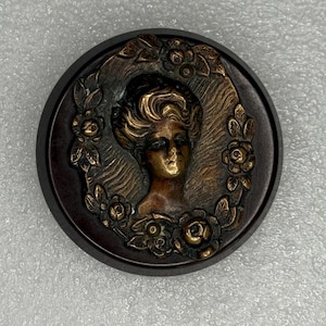May include: A round, dark brown button with a bronze-colored relief of a woman's profile surrounded by a floral wreath. The woman has detailed hair and facial features. The button's edge is a darker brown.