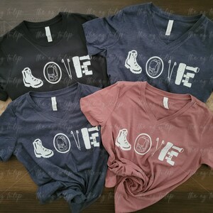 May include: Four different colored t-shirts with a white graphic design of hiking boots, a backpack, a compass, and a hiking stick. The graphic design spells out the word "LOVE".