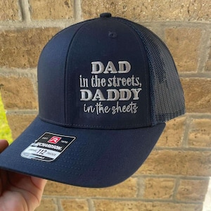 Puede incluir: Gorra de camionero azul marino con el texto bordado en plata "DAD in the streets, DADDY in the sheets". La gorra tiene una parte trasera de malla y una visera curva. Una etiqueta Richardson 112 Trucker es visible.