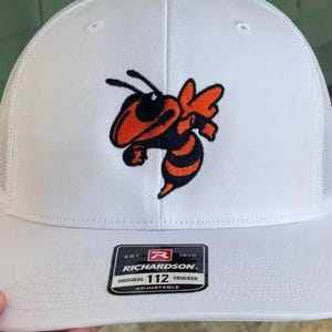 May include: White trucker hat with a mesh back and a curved brim. The hat features an embroidered orange and black hornet logo on the front. The Richardson patch is on the front, with the text "ORIGINAL 112 TRUCKER ADJUSTABLE."