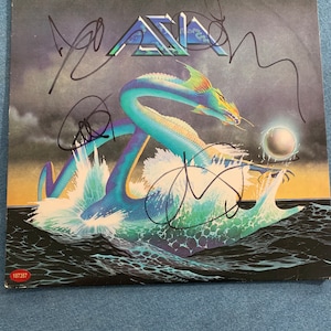 May include: A signed vinyl record album cover featuring a blue and green dragon emerging from the water. The album cover is for the band Asia and the title of the album is "Asia".