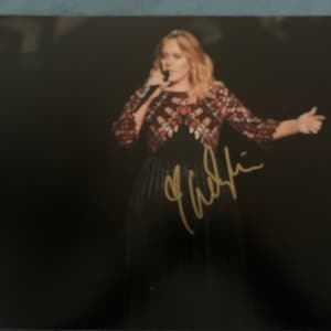 May include: A black and white photo of a woman singing into a microphone. She is wearing a black dress with a patterned top and has her hair pulled back. The photo is signed "Adele".