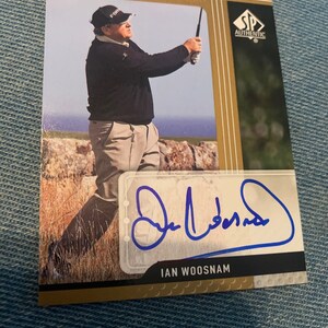 Puede incluir: Una tarjeta de intercambio de color dorado con una foto de un hombre con un suéter negro y pantalones caqui sosteniendo un palo de golf. La tarjeta tiene el texto "Sign Of The Times" e "IAN WOOSNAM". La tarjeta está firmada con tinta azul.