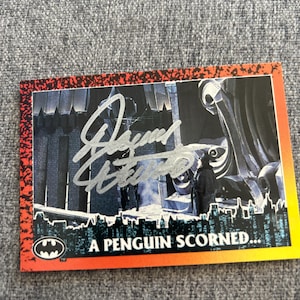 May include: A trading card featuring a scene from a Batman movie, with a signature in silver ink. The card has a red and orange border and the text "A PENGUIN SCORNED..." at the bottom, with the Batman logo.