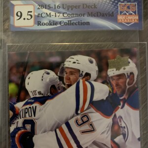 Puede incluir: Una tarjeta de hockey con Connor McDavid, un jugador de los Edmonton Oilers, que lleva una camiseta blanca con rayas azules y naranjas y el n&uacute;mero 97. La tarjeta est&aacute; calificada con 9,5 por Great Canadian Grading.