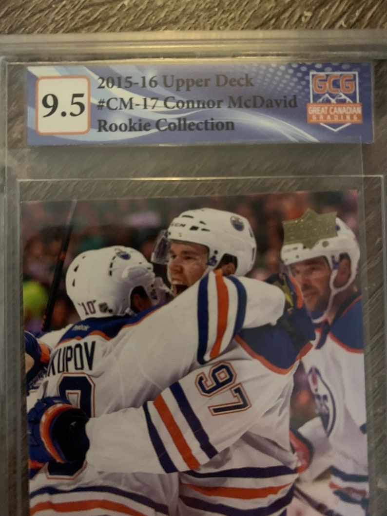 Puede incluir: Una tarjeta de hockey con Connor McDavid, un jugador de los Edmonton Oilers, que lleva una camiseta blanca con rayas azules y naranjas y el n&uacute;mero 97. La tarjeta est&aacute; calificada con 9,5 por Great Canadian Grading.