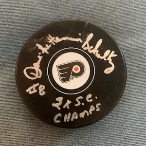 May include: Black hockey puck with a white Philadelphia Flyers logo and the signature "Eric Lindros" in white. The puck also has the text "'97 S.C. Champs" written in white.