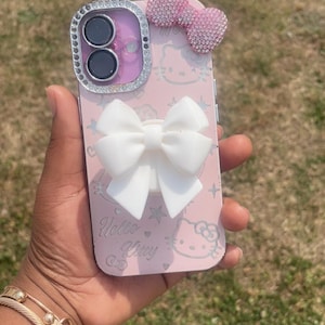 May include: Pink phone case featuring a Hello Kitty design, a white bow, and a sparkling pink bow. The case has a silver frame around the camera and the words "Hello Kitty" are visible on the case.