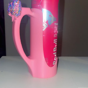 Pu&ograve; includere: Un portabottiglie rosa personalizzato con manico e fiocco tempestato di gioielli. Il supporto &egrave; progettato per contenere una lattina di Red Bull, visibile all'interno. La lattina &egrave; rosa con testo argentato e un logo.