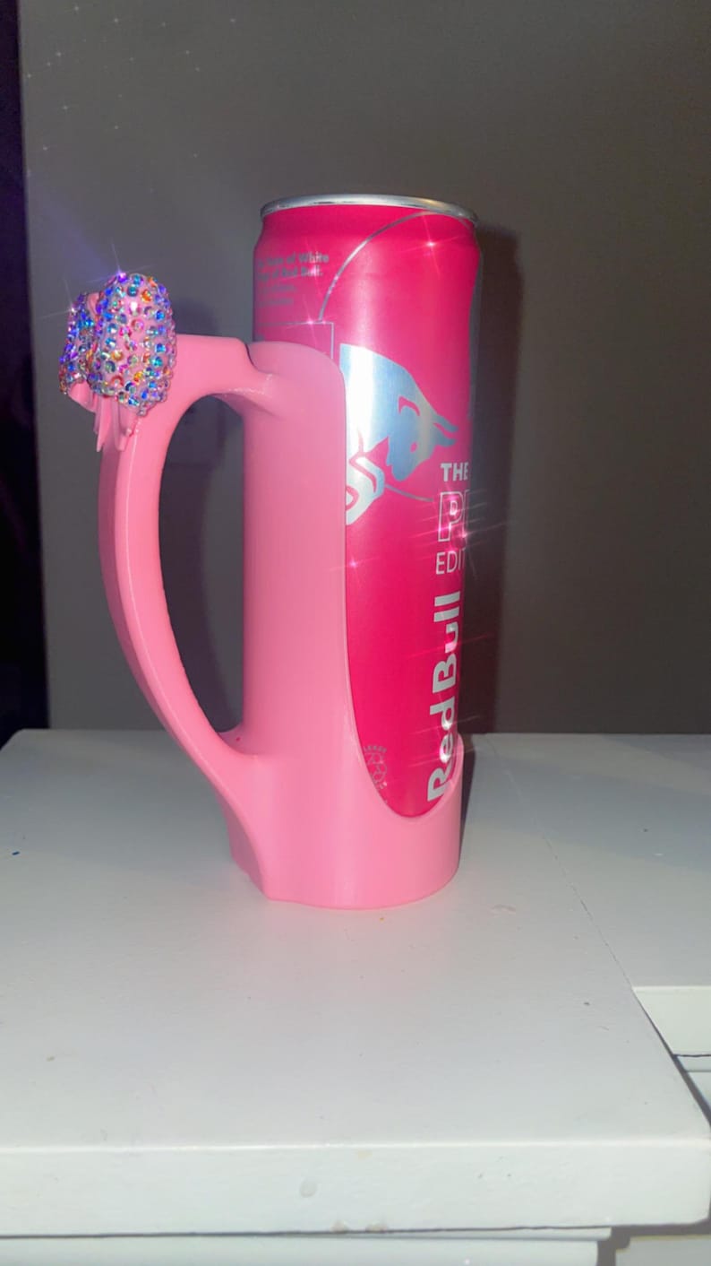 Pu&ograve; includere: Un portabottiglie rosa personalizzato con manico e fiocco tempestato di gioielli. Il supporto &egrave; progettato per contenere una lattina di Red Bull, visibile all'interno. La lattina &egrave; rosa con testo argentato e un logo.