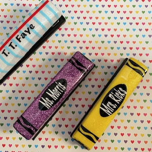 May include: Three colourful crayon-shaped teacher nameplates. The first is white with blue stripes and red accents, with the text "T.T. Faye". The second is purple with glitter and black accents, with the text "Ms. Morris". The third is yellow with glitter and black accents, with the text "Mrs. Ricks".