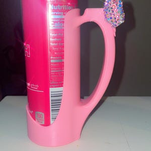 Pu&ograve; includere: Un porta lattina rosa con manico, che tiene una lattina di Red Bull Peach. Il manico &egrave; adornato da un gruppo di strass iridescenti. La lattina mostra informazioni nutrizionali e la parola "Peach".