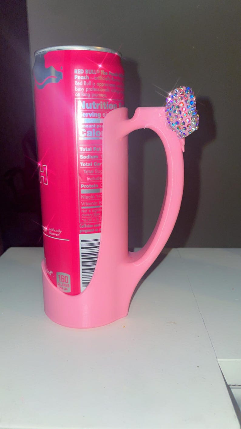 Pu&ograve; includere: Un porta lattina rosa con manico, che tiene una lattina di Red Bull Peach. Il manico &egrave; adornato da un gruppo di strass iridescenti. La lattina mostra informazioni nutrizionali e la parola "Peach".