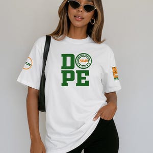 May include: White t-shirt with the word "DOPE" in large green block letters. Above the "PE" is a circular emblem with text. The shirt also has text on the sleeves. The person is wearing black shorts and sunglasses.