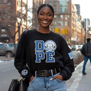 May include: Black sweatshirt with the word "DOPE" in white and blue block letters, with "Spelman College 1881" in a circular emblem. The sweatshirt has a crew neck and a small emblem on the sleeve. The person is wearing blue jeans.
