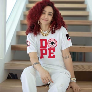 Peut inclure: T-shirt blanc avec le mot "DOPE" en lettres rouges. Le t-shirt présente également un logo et du texte de l'université Clark Atlanta. La personne porte un pantalon blanc et des bijoux dorés.