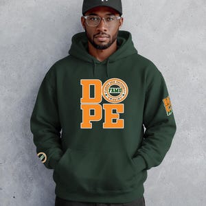 May include: Dark green hooded sweatshirt with the word "DOPE" in orange block letters. The letter "O" is replaced with a circular emblem that says "FAMU". The right sleeve has a matching emblem. The person is wearing a black baseball cap.