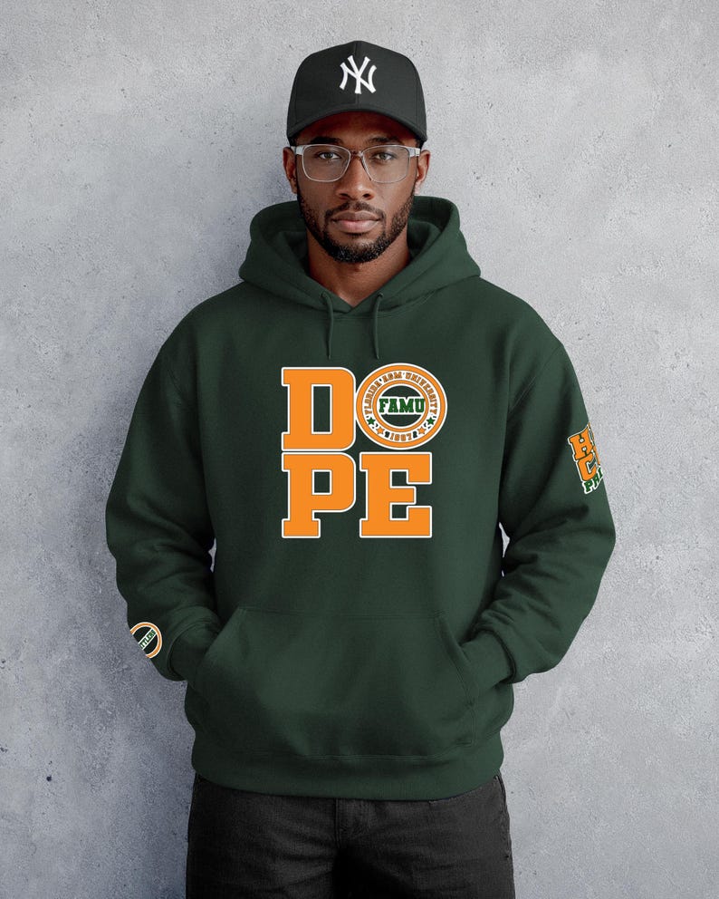 May include: Dark green hooded sweatshirt with the word "DOPE" in orange block letters. The letter "O" is replaced with a circular emblem that says "FAMU". The right sleeve has a matching emblem. The person is wearing a black baseball cap.