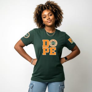 May include: A dark green t-shirt with the word "DOPE" in orange block letters. The shirt also features a circular logo and a patch on the sleeves. The person is wearing a gold chain and a watch.