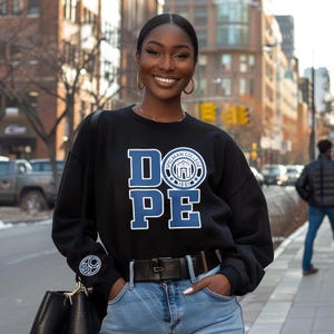 Może przedstawiać: Czarna bluza z napisem "DOPE" w niebieskich literach i emblematem Spelman College. Bluza ma długie rękawy i okrągły dekolt. Osoba ma na sobie niebieskie dżinsy i czarny pasek.